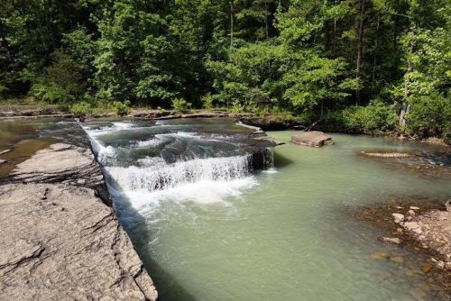 A serene waterfall cascades over rocky ledges into a calm, greenish pool, surrounded by lush trees.