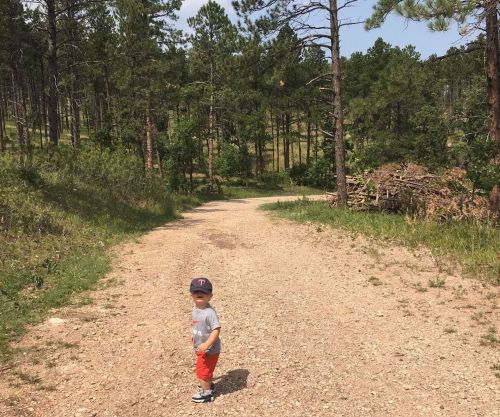 A young child in a gray shirt and cap stands on a dirt path surrounded by trees and greenery.