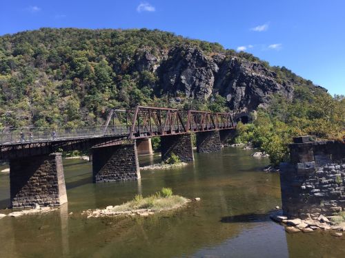 A rusted train bridge spans a calm river, surrounded by lush green hills and rocky cliffs under a clear blue sky.