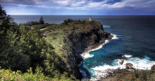 A scenic coastal view featuring a lighthouse on a rocky cliff, surrounded by lush greenery and crashing waves below.