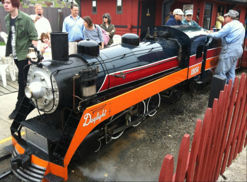 A vintage steam train with a shiny black and orange exterior, surrounded by a crowd of people at a station.