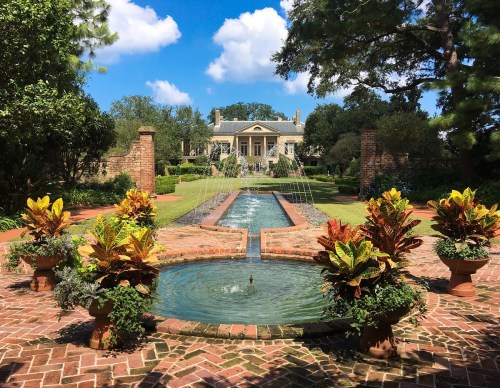 A lush garden with a fountain and colorful plants, leading to a grand mansion under a blue sky with fluffy clouds.