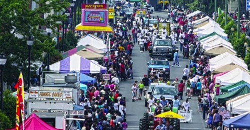 A bustling street fair with crowds, food stalls, and colorful tents lining the road under a clear sky.