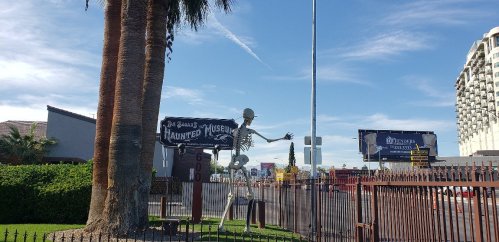 A large skeleton figure stands outside a haunted museum, with billboards and palm trees in the background.