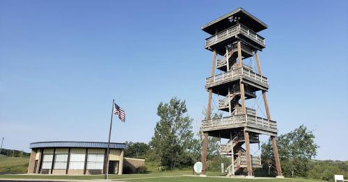 A tall wooden observation tower next to a modern building, with a flagpole displaying the American flag.