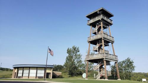 A tall wooden observation tower next to a modern building, with a flagpole and trees under a clear blue sky.