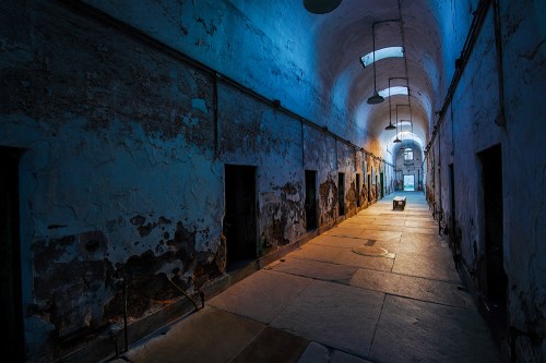 A dimly lit, abandoned prison corridor with peeling walls and empty cell doors on either side.