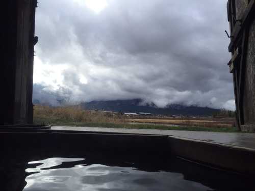 View from a rustic structure overlooking a calm water surface, with dramatic clouds and mountains in the background.