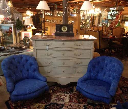 A vintage dresser with four drawers, flanked by two blue tufted chairs, set in a cozy, eclectic furniture store.