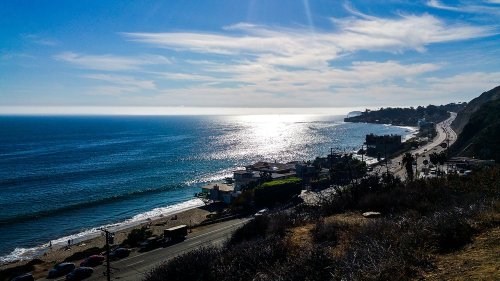A scenic view of the ocean with a coastline, reflecting sunlight, and a road lined with buildings and palm trees.