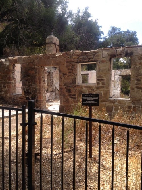Ruins of a stone building with windows, surrounded by overgrown vegetation and a fence, labeled "Katherine House."