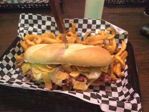 A large sandwich with meat, cheese, and toppings, served with curly fries on a checkered paper-lined tray.