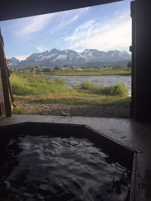 View from a rustic cabin showing a hot tub with mountains and a lake in the background under a clear blue sky.