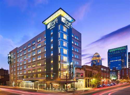 Aloft hotel building at dusk, featuring modern architecture and illuminated signage, with city skyline in the background.