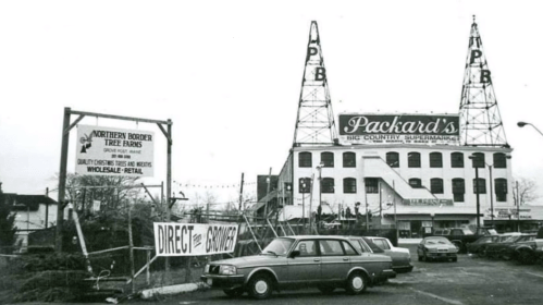 Black and white photo of Packard's Big Country Supermarket with vintage cars parked outside and signage visible.