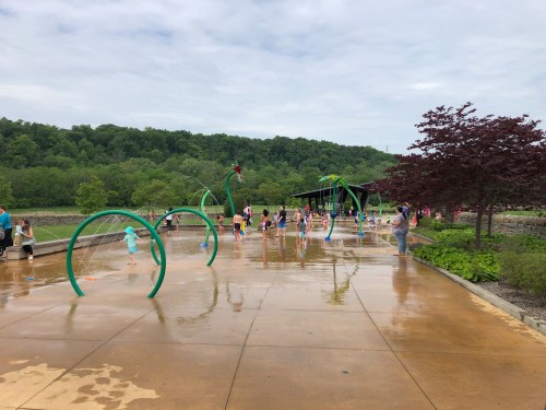 Children play in a splash pad area with water features, surrounded by greenery and a pavilion in the background.