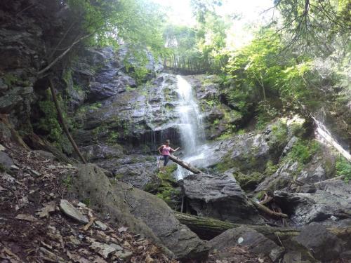 A person climbs over rocks near a waterfall surrounded by lush greenery and sunlight filtering through the trees.