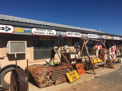 A rustic BBQ shop with vintage signs, an old truck hood, and a swing, set against a clear blue sky.
