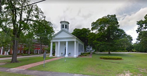 A white building with a steeple surrounded by trees and green grass, set against a cloudy sky.
