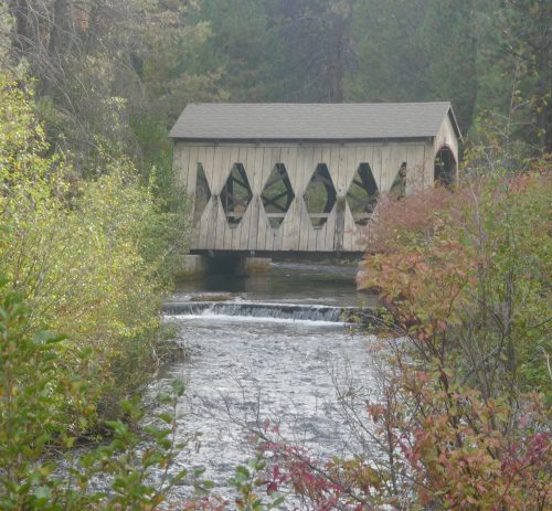 A wooden covered bridge spans a serene stream, surrounded by lush greenery and autumn foliage.