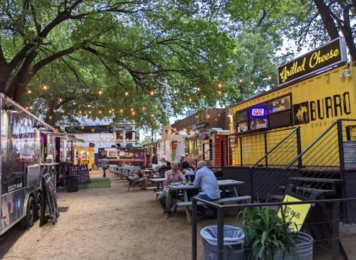 A vibrant food truck park with string lights, picnic tables, and various food vendors under lush green trees.