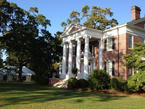 A grand brick mansion with white columns, surrounded by lush greenery and a large tent in the background.