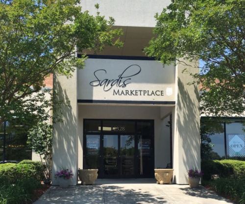 Entrance of Sardis Marketplace with a sign, surrounded by greenery and flower pots.