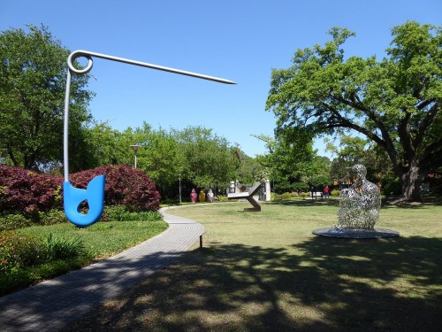 A large blue safety pin sculpture and a wire figure in a park with trees and visitors in the background.