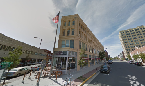 Historic building with an American flag, outdoor seating, and parked cars on a city street.