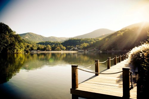 A serene lake view with a wooden dock, surrounded by lush mountains and trees under a warm, golden sunlight.