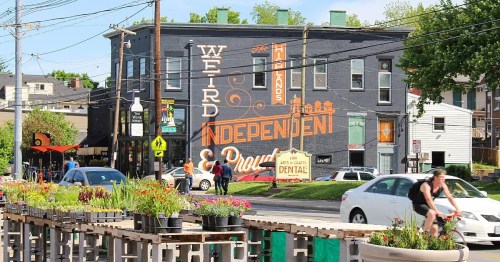 A vibrant street scene featuring a mural that reads "Weird, Independent & Proud" on a building, with flowers and cyclists nearby.