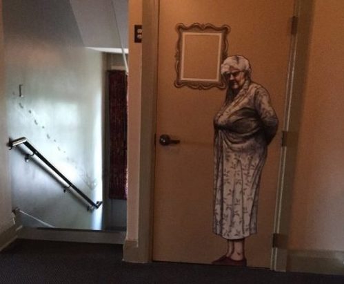 A door with a mural of an elderly woman in a robe stands in a dimly lit hallway with stairs in the background.