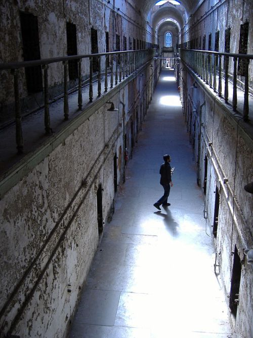 A dimly lit prison corridor with peeling walls, featuring a solitary figure walking in the light.