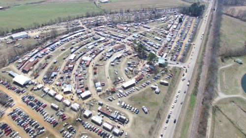 Aerial view of a large outdoor market with numerous stalls, vehicles, and people in a rural setting.