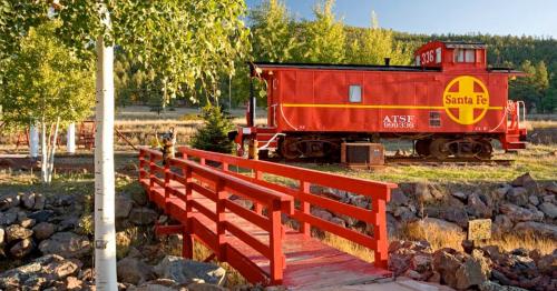 A bright red Santa Fe caboose next to a wooden bridge, surrounded by trees and rocky terrain.