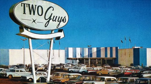 A vintage storefront sign for "Two Guys" with a parking lot full of classic cars under a clear blue sky.