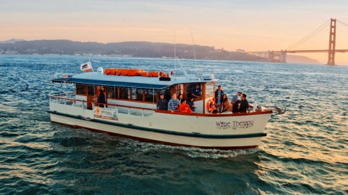 A boat named "Wine Therapy" with passengers cruising on water near the Golden Gate Bridge at sunset.