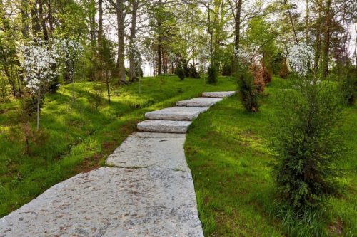 A stone pathway winds through a lush green landscape with trees and flowering plants.
