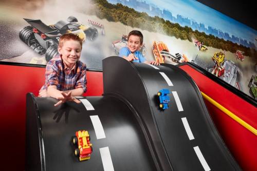 Two boys play with toy cars on a curved track, smiling against a colorful backdrop of vehicles and a cityscape.