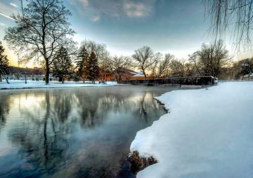 A serene winter landscape featuring a snowy riverbank, trees, and a building reflected in the calm water at sunset.