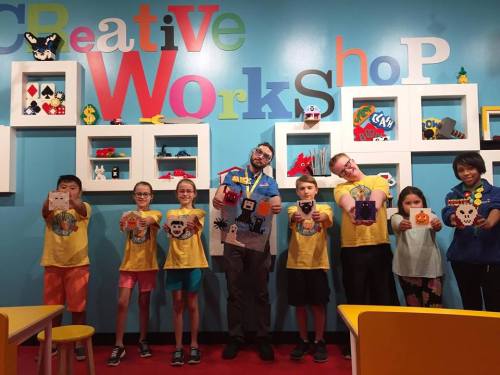 A group of children in yellow shirts hold up their crafts in front of a colorful workshop wall with shelves of art.