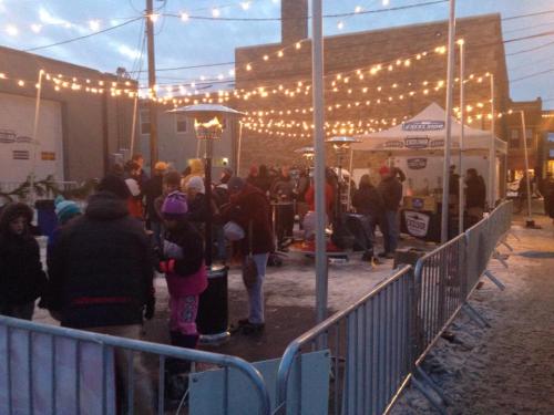 A festive outdoor scene with people enjoying activities under string lights, surrounded by barriers and a vendor tent.