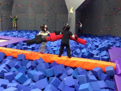 Two children in helmets play on a balance beam over a pit filled with blue foam blocks, while others climb nearby.