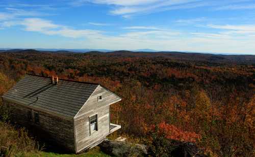 A rustic cabin overlooks a vibrant autumn landscape filled with colorful trees and distant mountains under a blue sky.