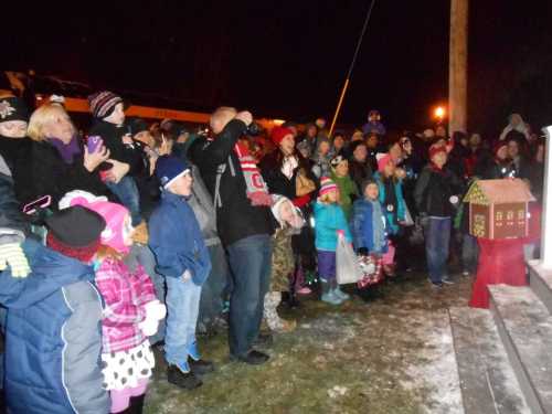 A crowd of children and adults in winter clothing gathers at night, watching a festive event near a small decorated house.