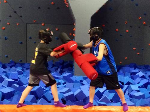 Two kids in helmets sparring with padded weapons over a blue foam pit in a colorful indoor play area.