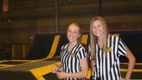 Two smiling female referees in striped shirts stand in a trampoline park, with trampolines visible in the background.