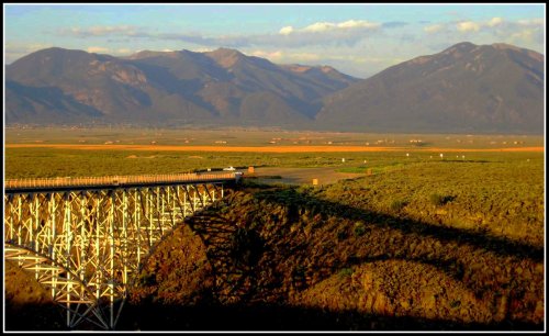A scenic view of a bridge casting a shadow over green fields, with mountains in the background under a clear sky.