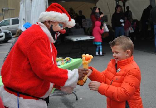 A clown in a Santa suit hands a balloon animal to a smiling boy in an orange jacket at a festive outdoor event.