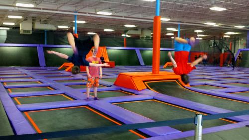 Two kids perform flips on trampolines in a colorful indoor trampoline park, with others playing in the background.